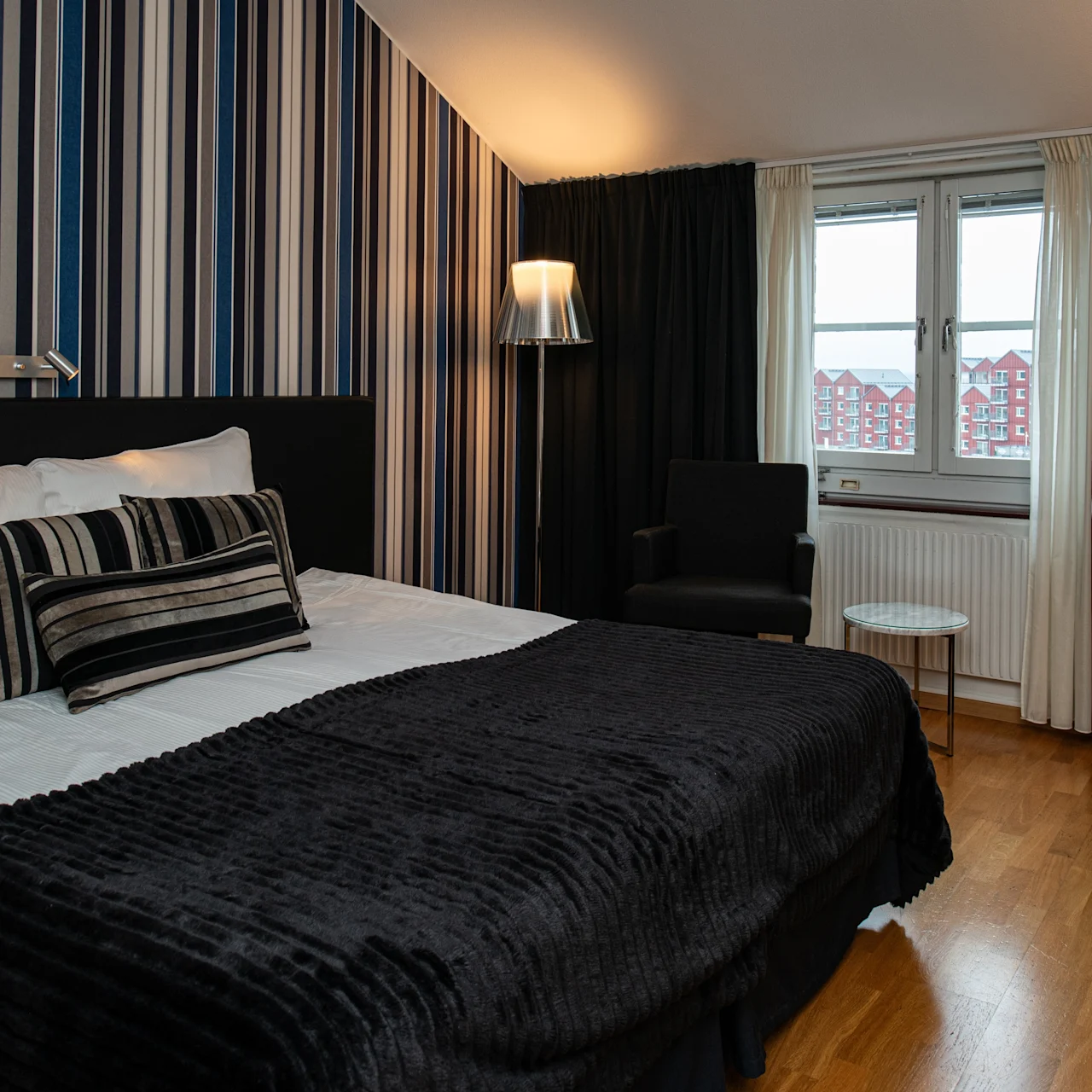 En pent oppredd seng med svart-hvite stripete puter og et mørkt teppe står i et moderne soverom. En stol og et lite bord står ved et vindu, med stripete tapet.