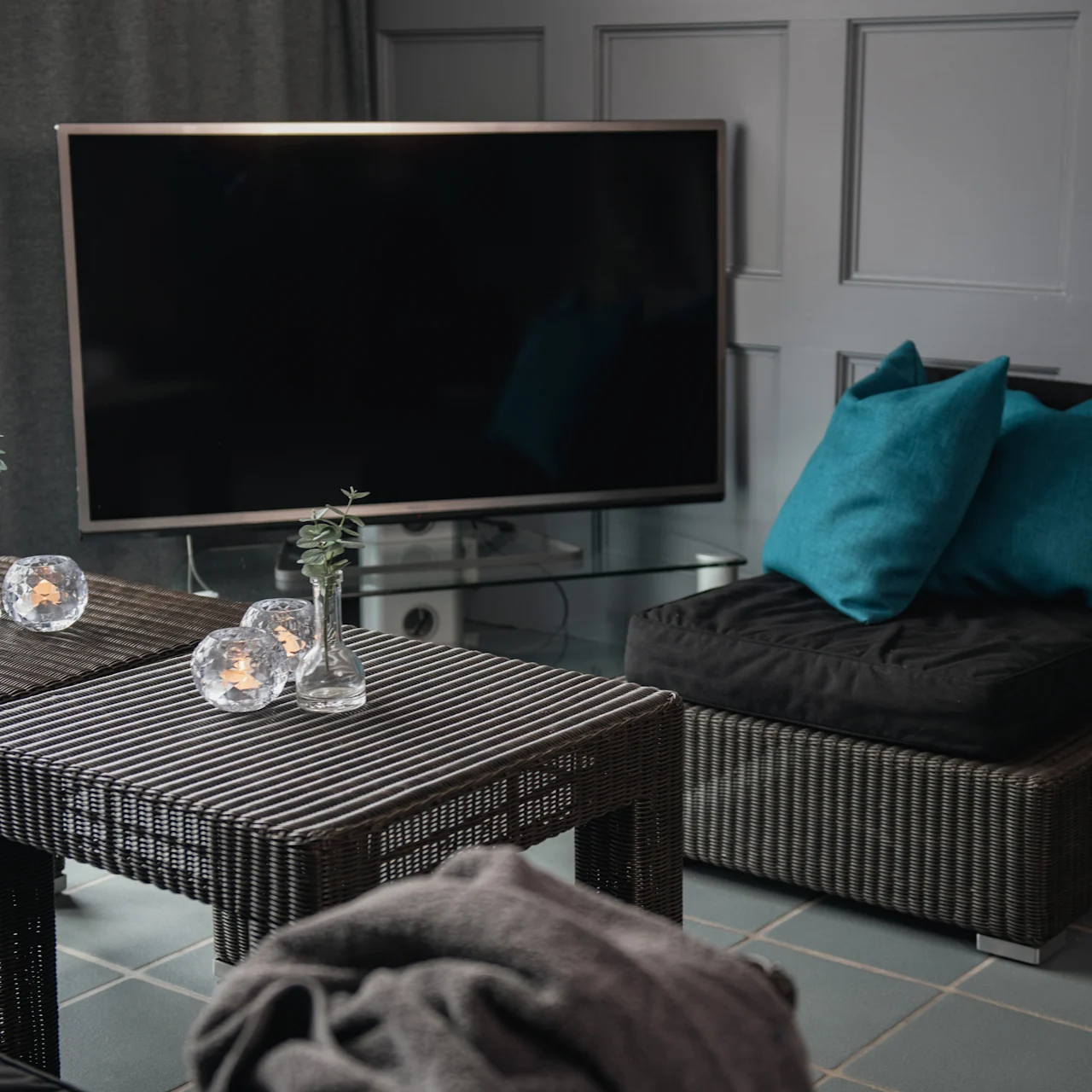 A flat-screen TV is off in a modern living room with a wicker table, small vases, gray sofas, and teal cushions.
