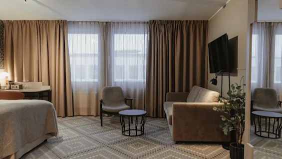 Ett modernt och inbjudande hotellrum med en bekväm säng, en sittgrupp och stora fönster med gardiner.