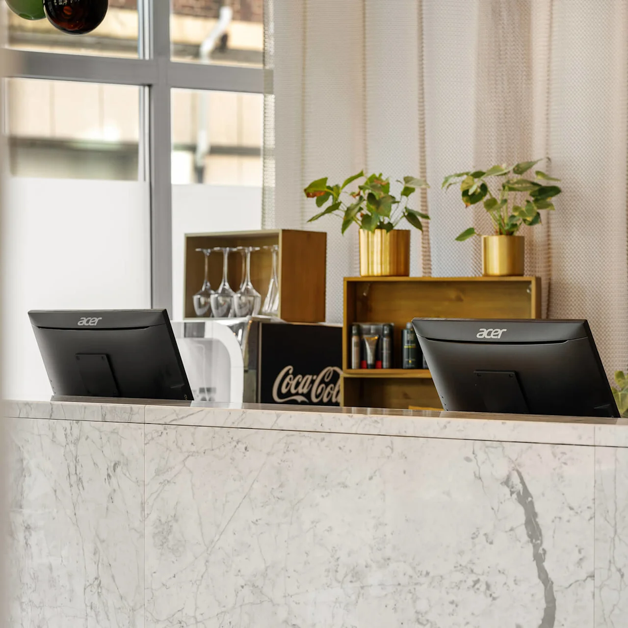 To computerskærme står på en marmor-reception. Bag dem er planter i guldfarvede potter, glas og et "Coca-Cola"-skilt på en hylde med et vindue i baggrunden.