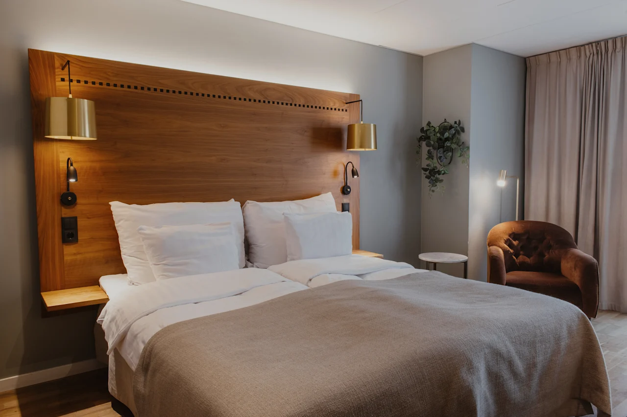 Et koselig hotellrom med en stor seng, sengegavl i tre og varm belysning, med en komfortabel lenestol og et snev av grønt.