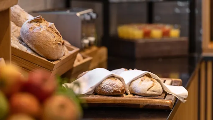 Leipiä lepää puisella laudalla, osittain valkoisen liinan peitossa, kiiltävällä keittiötasolla. Korillinen hedelmiä on epätarkka etualalla.