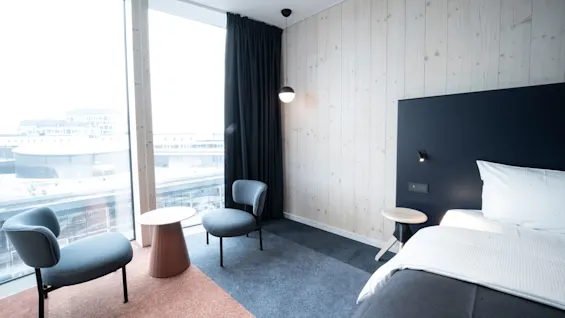 Modern hotel room with a bed, two chairs, and tables, framed by a large window and dark curtains. Light wood wall panels create a minimalist, airy environment.