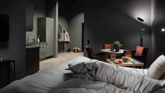 A bed with rumpled sheets sits in a dark room, facing a small kitchen area and a dining table with chairs in the background.