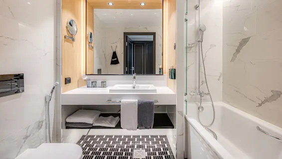 Modern bathroom with a large mirror above a sink; towels hang below. To the right, a bathtub with a showerhead; marble walls create an elegant atmosphere.