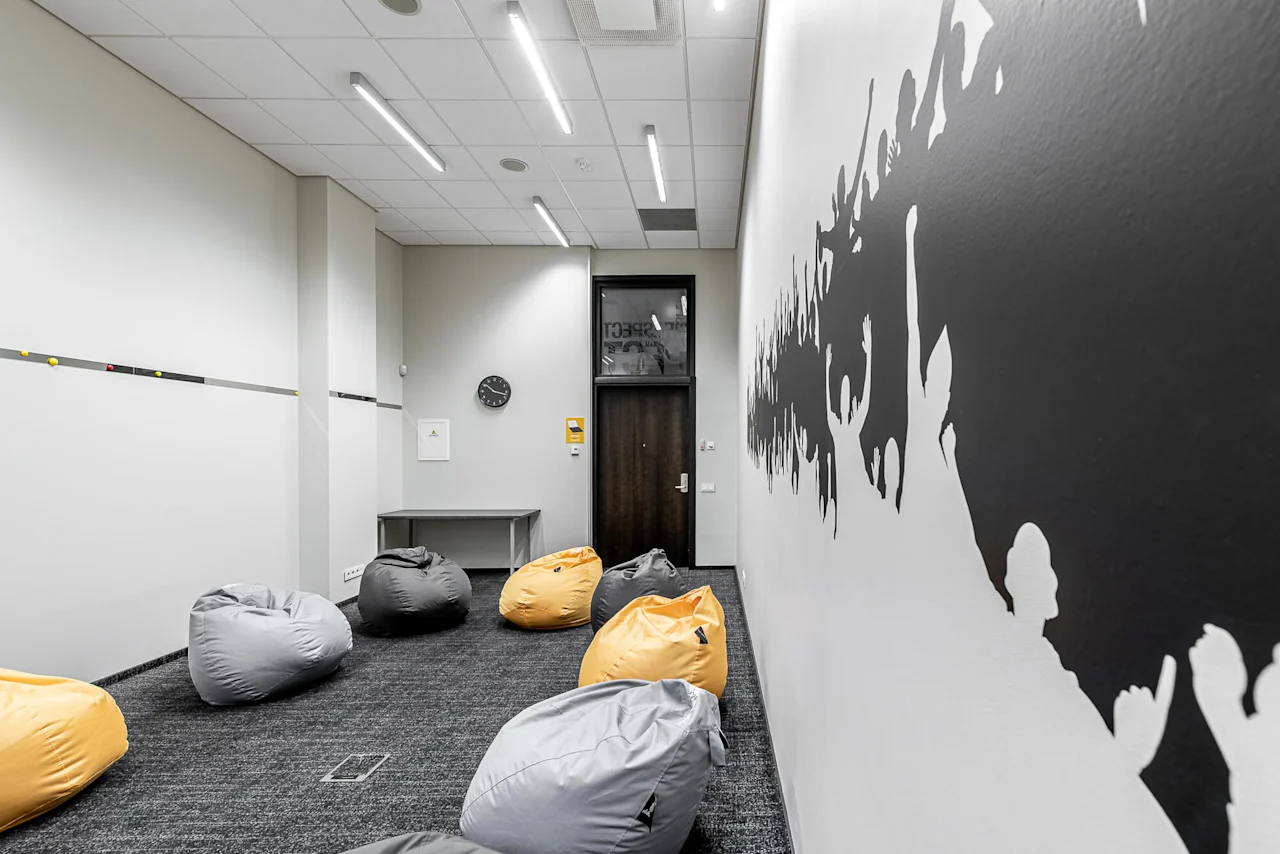 Bean bags rest on a carpeted floor in a minimalist room. A black silhouette mural decorates the wall, and a clock hangs above a small table. Text includes "RESPECT."