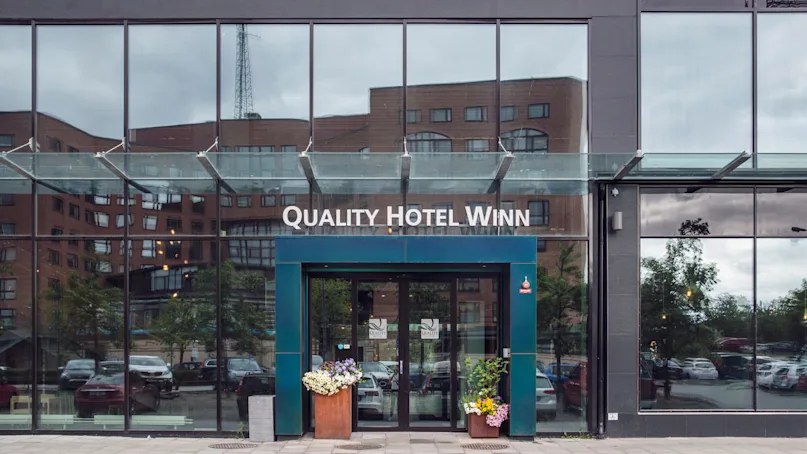Quality Hotel Winn entrance features automatic glass doors, flanked by flower arrangements in a modern urban setting, with reflections of nearby buildings.  