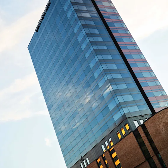 En høy glasskyskraper reflekterer himmelen, som tårner over en murbase. Teksten lyder "CLARION COLLECTION FRIMURARE". Bygningen er mot en delvis skyet himmel.