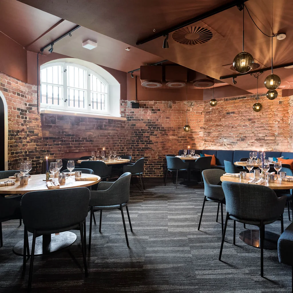 Borden är dukade med glas och bestick; omgiven av stolar och soffor; i en mysig, svagt upplyst restaurang med exponerade tegelväggar och moderna hängande lampor.