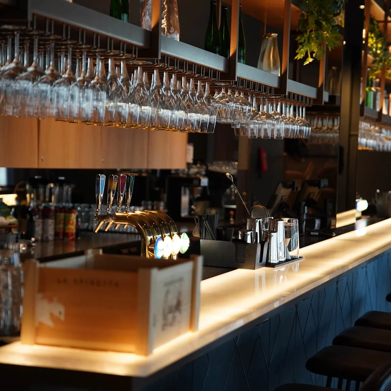 Bardisk pyntet med hængende glas, en ølhane og cocktailshakere, placeret i en svagt oplyst, elegant indstilling. En trækasse mærket "La Spinetta" står på disken.