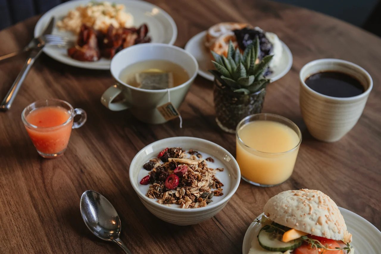 Home Hotel Post: En utsökt frukostbuffé på ett träbord med ägg, bacon, granola, juicer, kaffe och en smörgås.