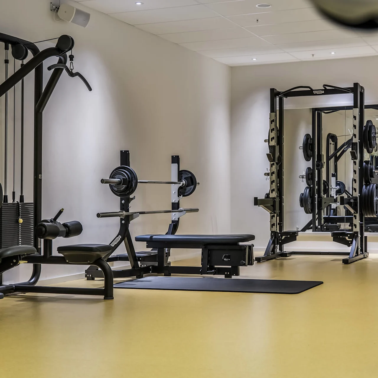 Weightlifting equipment, including barbells and a bench press, stands unused in a well-lit gym with mirrors, a yellow floor, and a visible exercise ball branded with "TECHNOGYM".