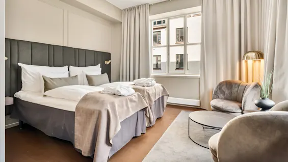 Et pænt hotelværelse med en stor seng og hvide puder ved siden af et vindue. To plysstole og et rundt bord supplerer den blødt oplyste, moderne indretning.