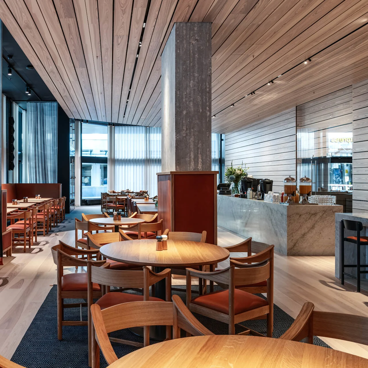 Elegant restaurant featuring wooden tables and chairs, neatly arranged for dining. Soft lighting filters through sheer curtains, creating a serene, modern atmosphere with a visible counter in the background.