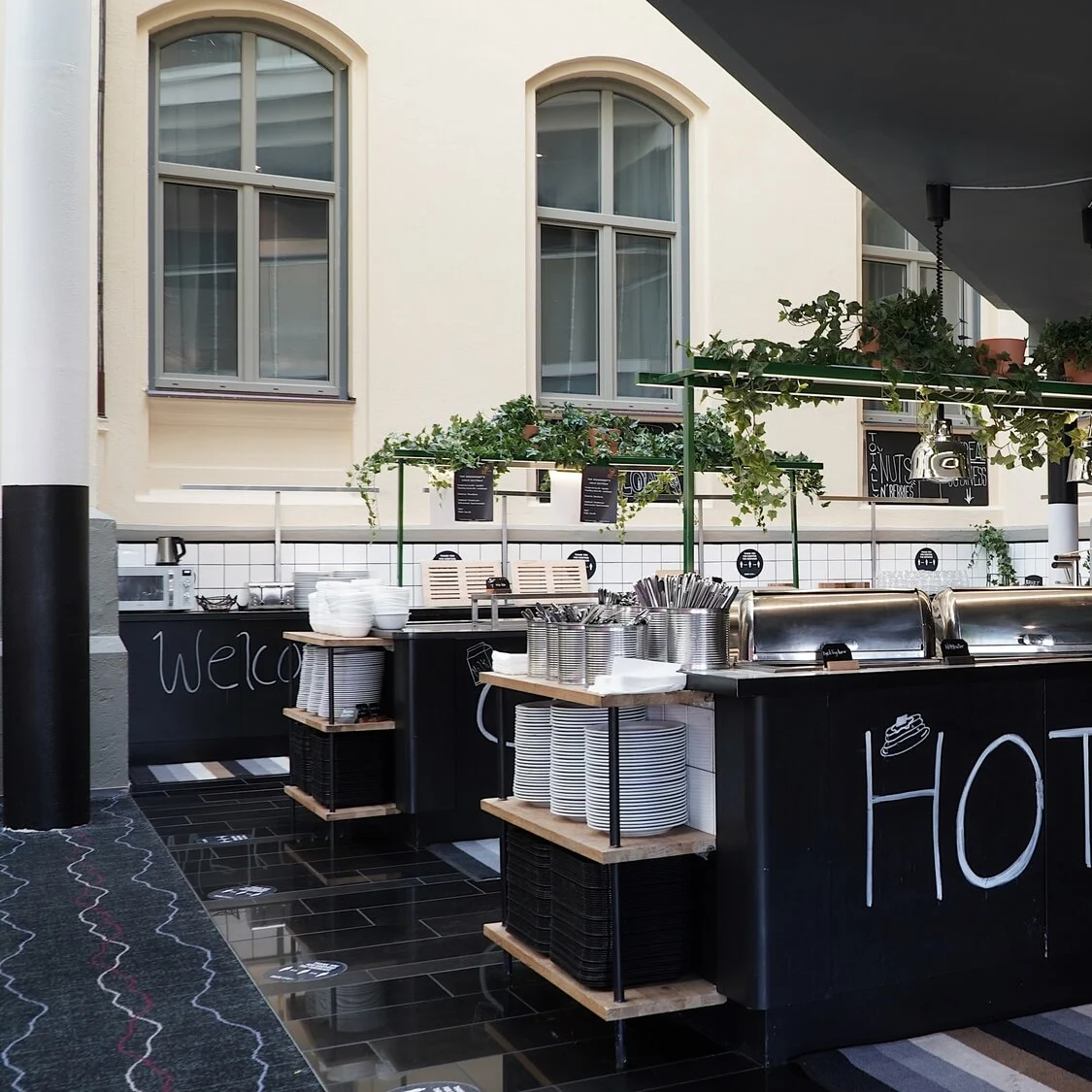 A buffet station with the words "HOT STUFF" on its front, stocked with plates and serving utensils, in a modern interior space with large windows.