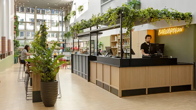 Baaritiski yläpuolella vihreää, työntekijä kassalla ja neonkyltti "BARCEPTION". Sisäympäristö, jossa kasveja ja istuma-alueita taustalla.