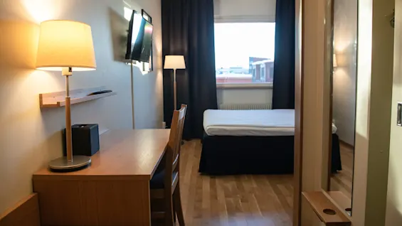 A small hotel room has a bed facing a window with curtains, a wall-mounted TV, and a wooden desk with a lamp and chair on a wooden floor.