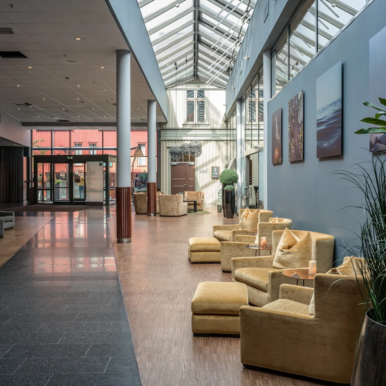 Bekväma beige fåtöljer arrangerade i en rymlig lobby, omgivna av höga växter och modern konst, under ett glastak, som skapar en inbjudande och luftig sittplats.