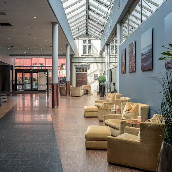 Behagelige beige lenestoler plassert i en romslig lobby, omgitt av høye planter og moderne kunstverk, under et glasstak, som gir et innbydende og luftig sitteområde.