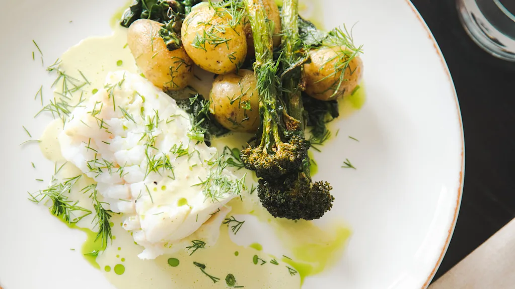 Seared white fish with potatoes and broccoli topped with fresh dill on a white plate, surrounded by a light green sauce, garnished with green herbs.