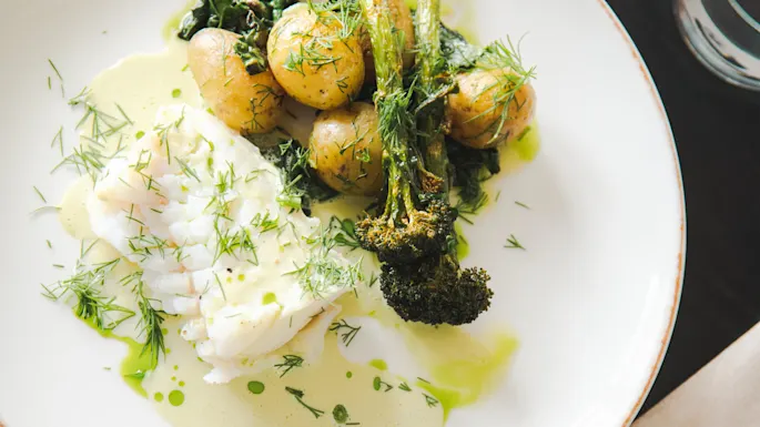 Panert hvit fisk med poteter og brokkoli toppet med fersk dill på en hvit tallerken, omgitt av en lysegrønn saus, pyntet med grønne urter.