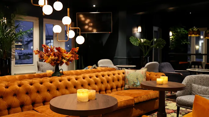 A tufted orange sofa with lit candles on round tables, set in a dimly lit, elegant lounge featuring modern spherical chandeliers, decorative pillows, and plants near large windows.