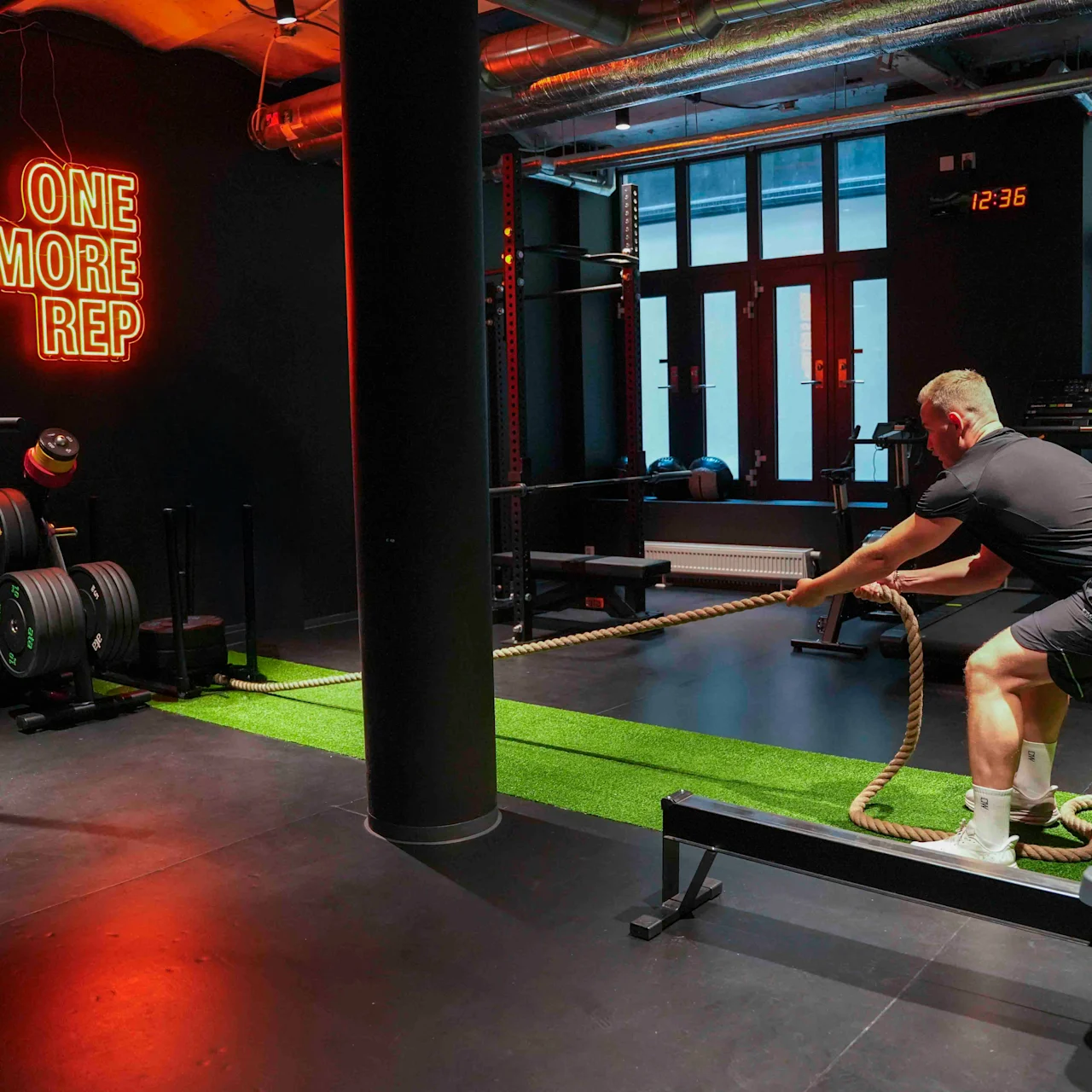 En person trekker i et tungt tau innendørs, omgitt av treningsutstyr; neonskiltet sier "ONE MORE REP" og veggklokken viser 12:36.