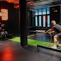 En person trekker i et tungt tau innendørs, omgitt av treningsutstyr; neonskiltet sier "ONE MORE REP" og veggklokken viser 12:36.