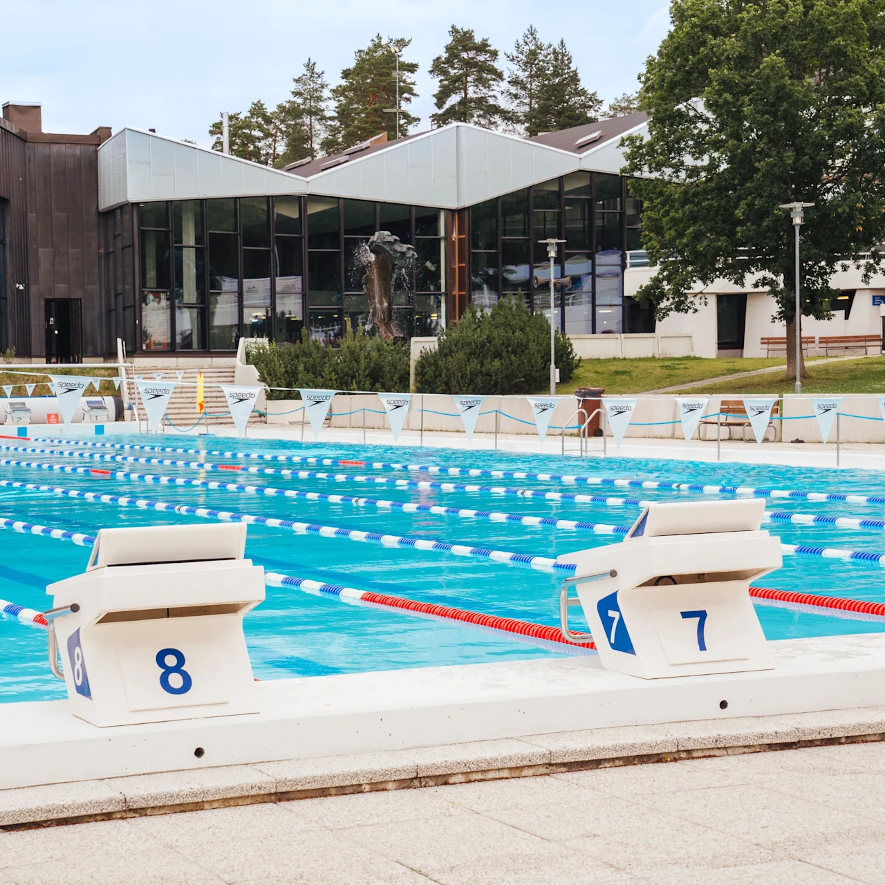 A swimming pool with blue lanes numbered 5, 6, 7, and 8, is surrounded by a modern building and trees.  