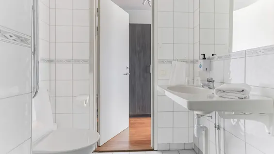 A bright and clean bathroom featuring white tiles, a toilet, a sink with fresh towels, and an open doorway.