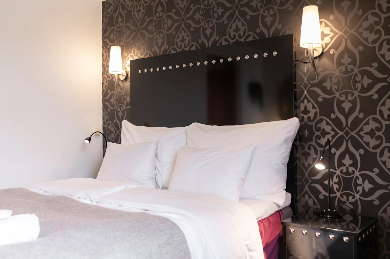 Viihtyisä hotellihuone, jossa on sänky valkoisilla liinavaatteilla, tumma kuvioitu sängynpääty ja lämmin, kutsuva valaistus.