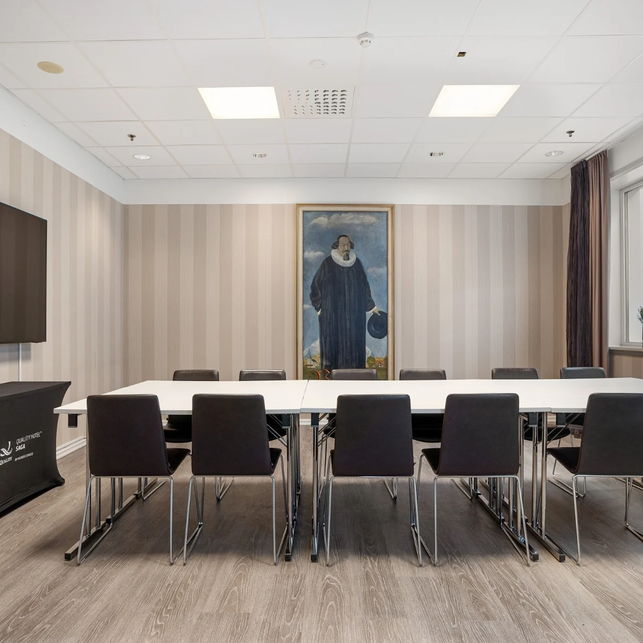 A large table surrounded by black chairs in a conference room. A portrait hangs on the wall. A flat-screen TV is mounted on the left. Beige-striped walls add a formal touch.