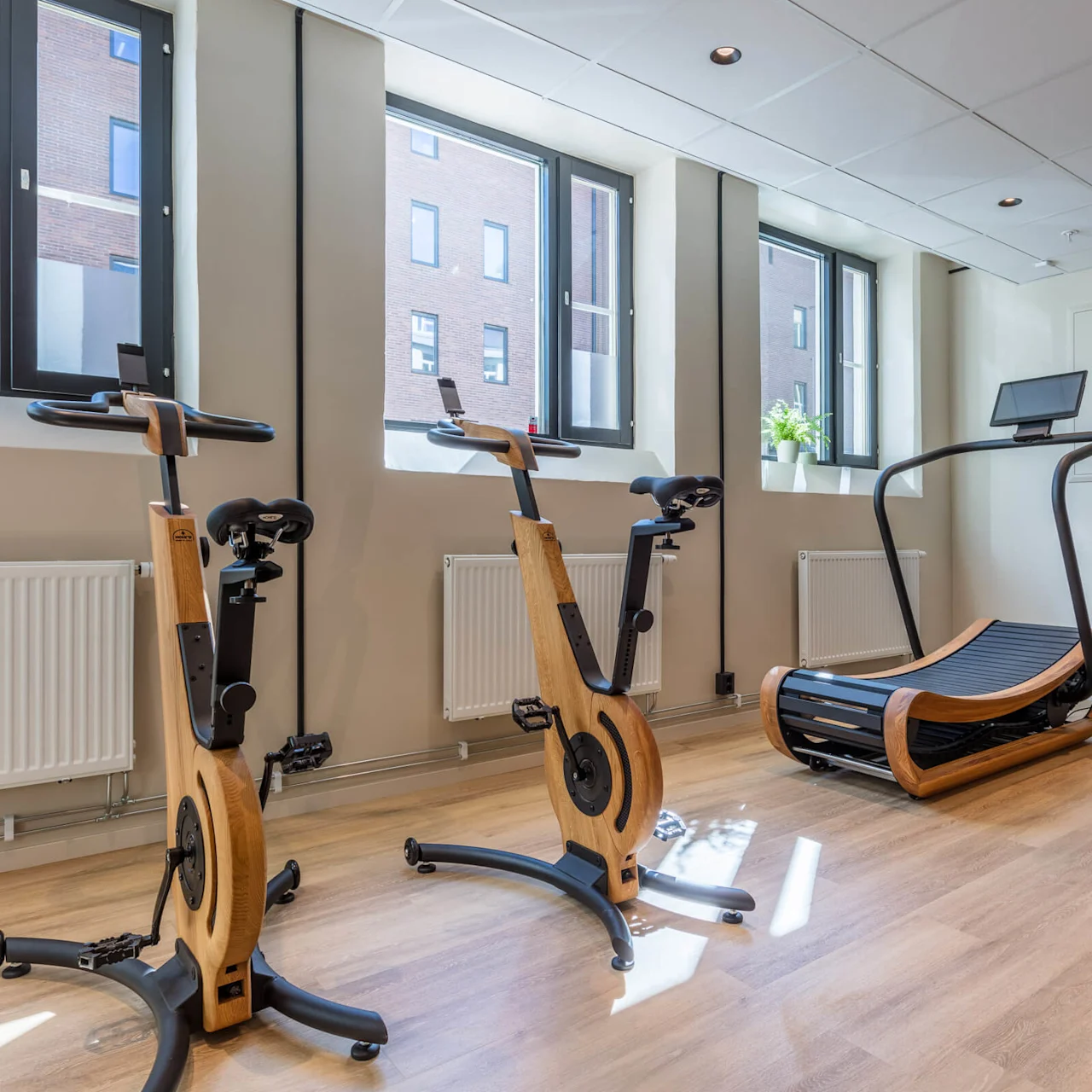 Exercise bikes and a rowing machine in a bright, modern gym with wooden floors and windows.