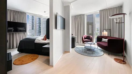 Bedroom with a modern bed, facing a flat-screen TV; adjacent living area features maroon seating, a floor lamp, and round tables. Large windows with city skyscrapers visible outside.