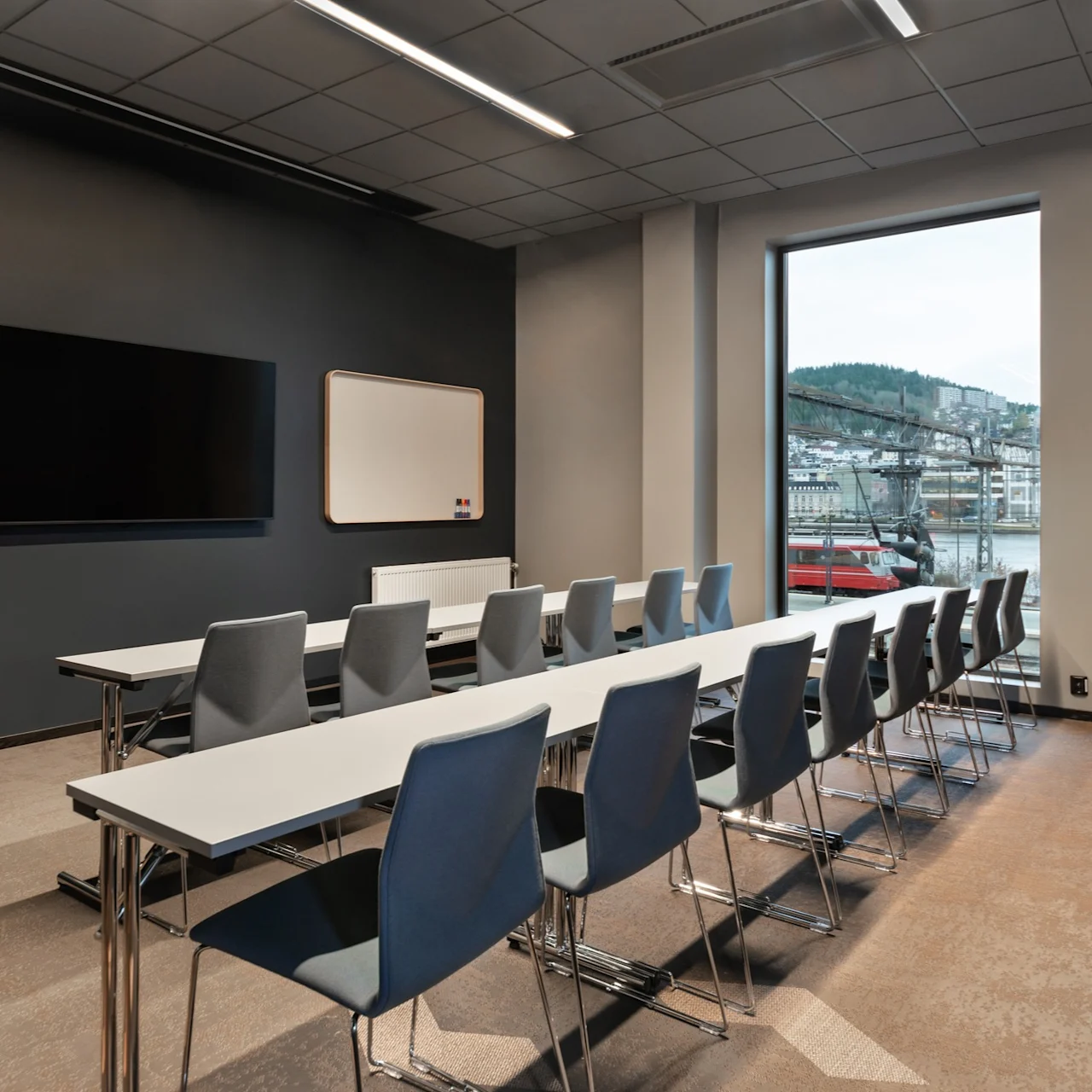 En rekke tomme stoler vender mot en stor skjerm og whiteboard i et moderne konferanserom med store vinduer og utsikt over bygninger og åser utenfor.