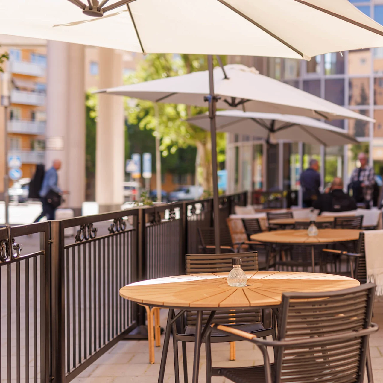 An outdoor cafe seating area with empty tables and sun umbrellas on a sunny day, people in the background.