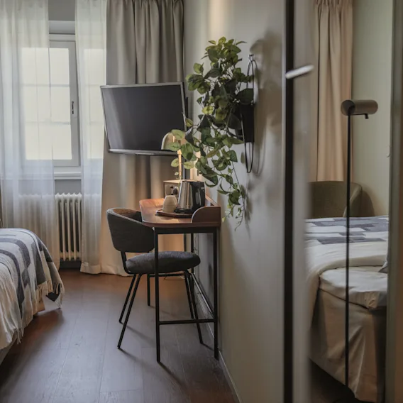 Et hyggeligt hotelværelse med en seng, et lille skrivebord med en elkedel, et vægmonteret TV og en plante.
