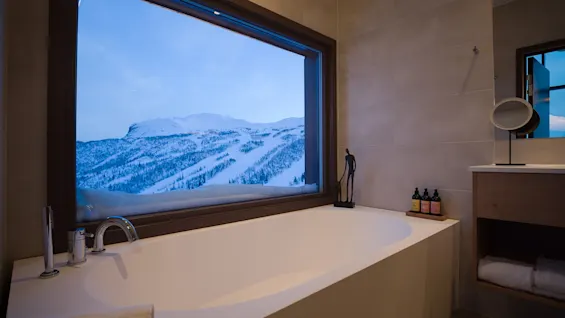 A white bathtub sits under a large window, offering a view of snow-covered mountains. Nearby, shelves hold towels, and bottles are placed on a tray.