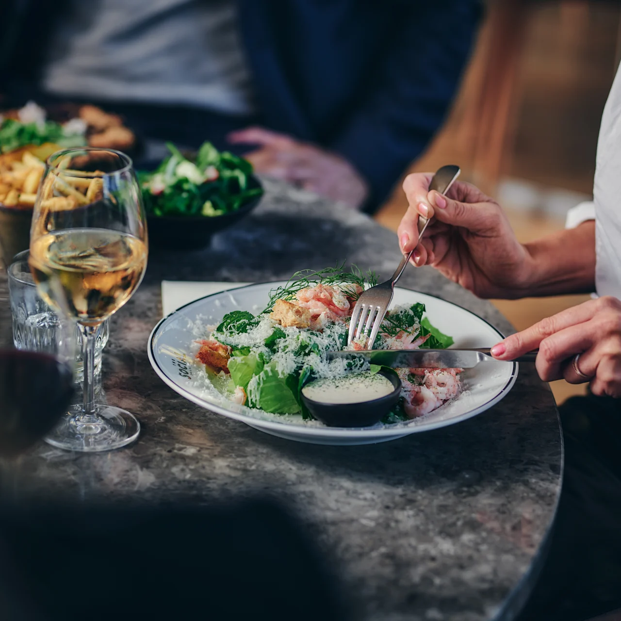 En person äter en skaldjurssallad med bestick vid ett restaurangbord, tillsammans med ett glas vitt vin, ett glas vatten och andra middagsgäster i bakgrunden.