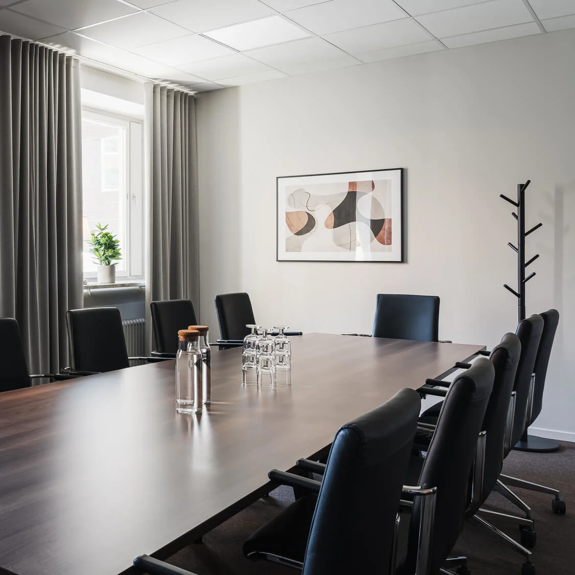 Ett långt konferensbord med läderstolar är dukat med vattenflaskor och glas, beläget i ett modernt kontor med fönster, gardiner och abstrakt väggkonst.