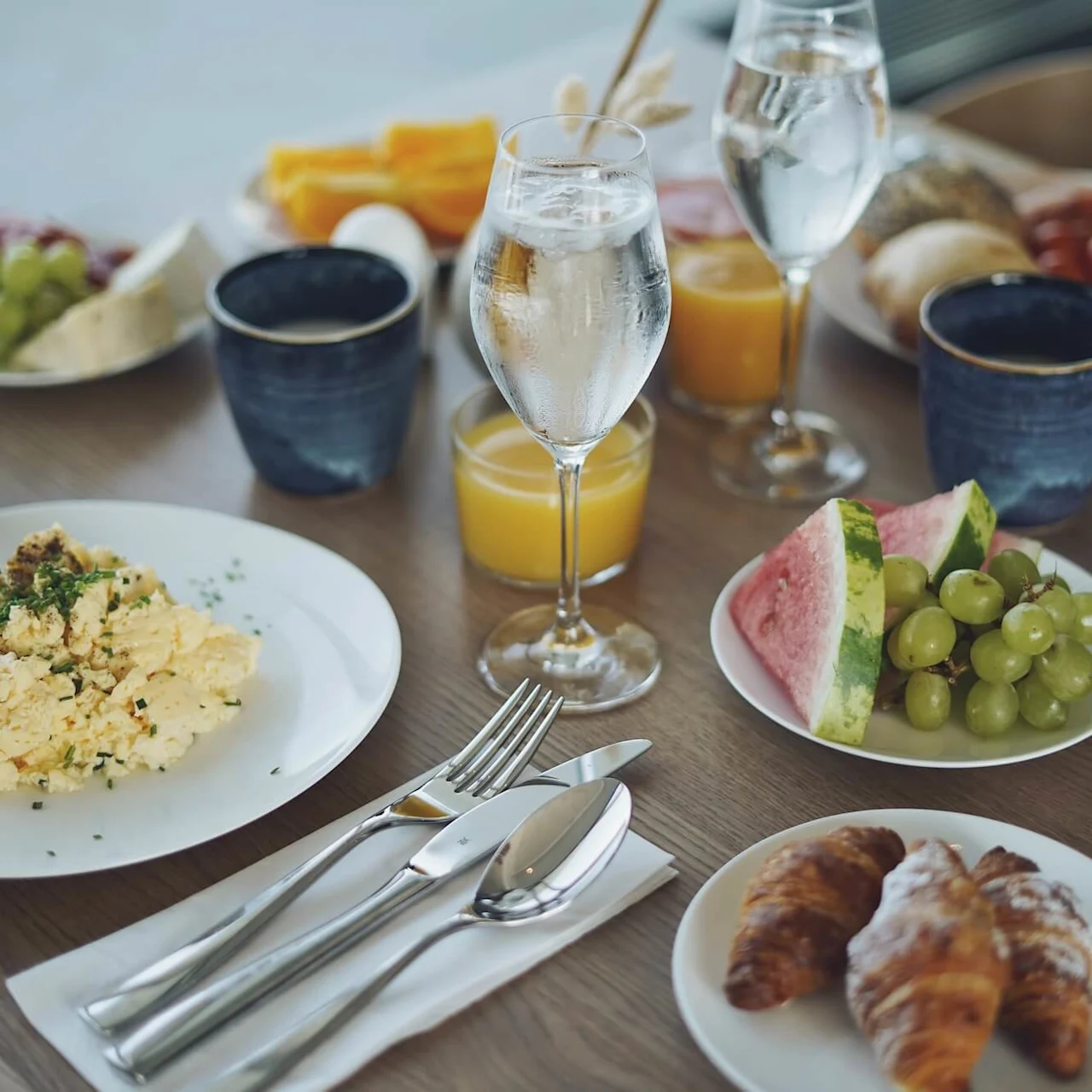Eggerøre ligger på en hvit tallerken, omgitt av fersk frukt, croissanter og drikke på et trebord.