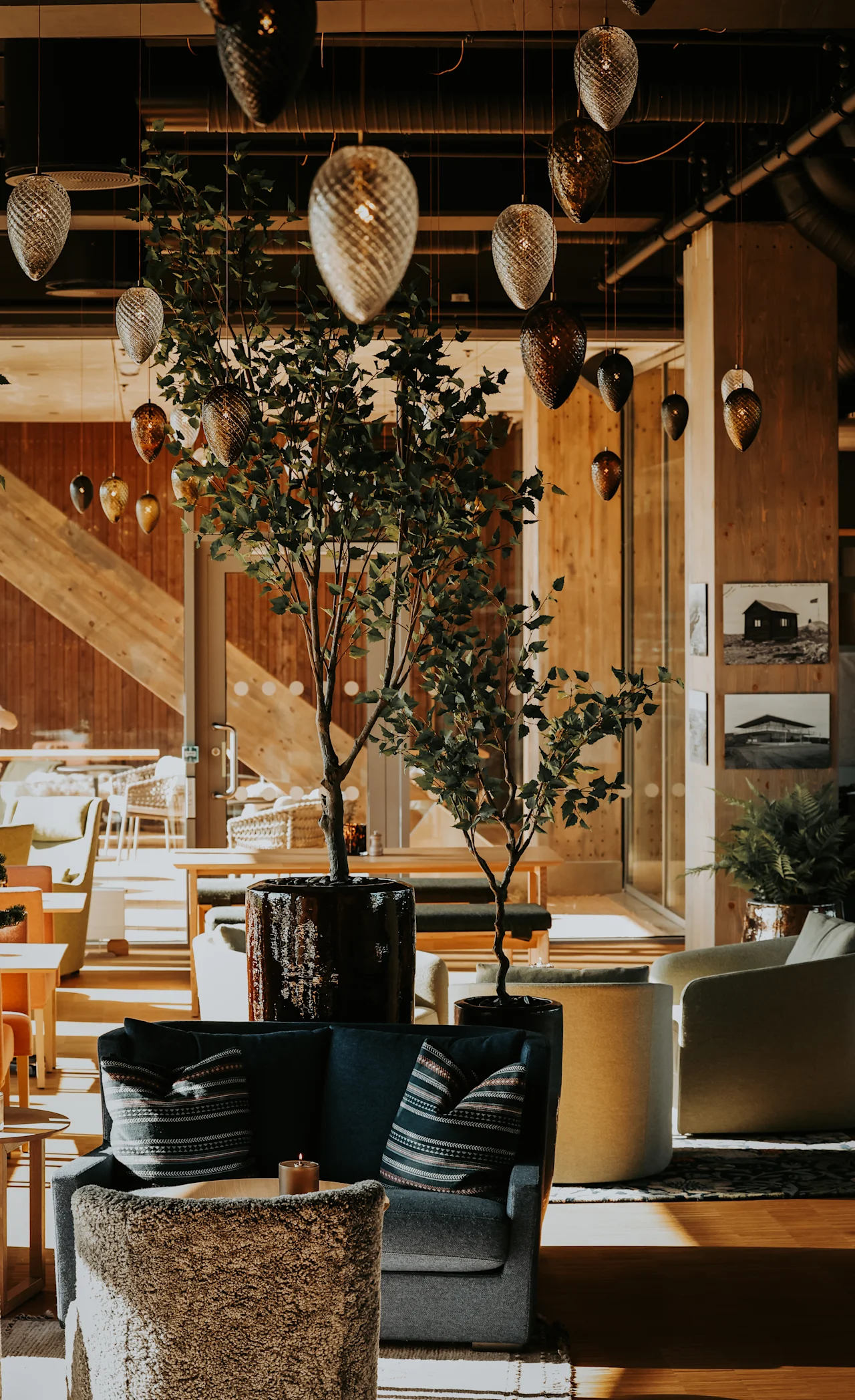 En hyggelig lounge har en mørk sofa med stribede puder, omgivet af løvrige planter og varme trævægge. Elegante pendellamper hænger fra loftet og oplyser rummet.