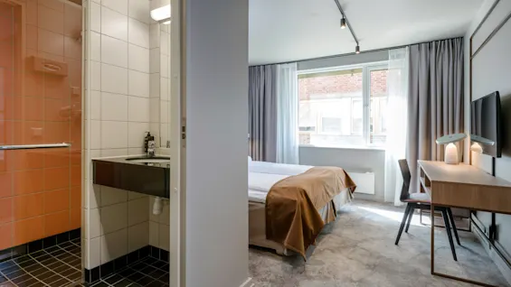 Et hotelværelse med en seng og skrivebord nær et stort vindue; ved siden af er der et badeværelse med vask og bruser. Rummet er moderne og godt oplyst.