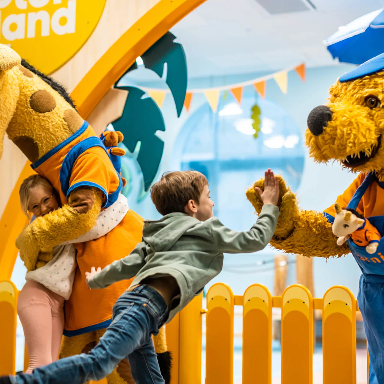 En giraf-maskot omfavner et barn, mens en bjørne-maskot giver high five til et andet barn foran en farverig legeplads, med et skilt der siger "Sodor Land."