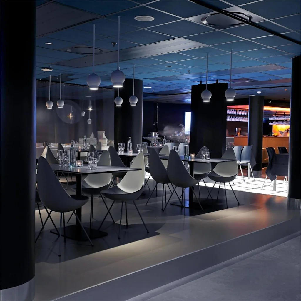 Modern dining area with styled tables and chairs, spherical hanging lights, and a bar in the background with ambient lighting.