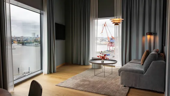 Ett mysigt rum med en grå soffa, ett runt bord med röda blommor och stora fönster. Utanför syns en kran mot en industriell vattensilhuett.