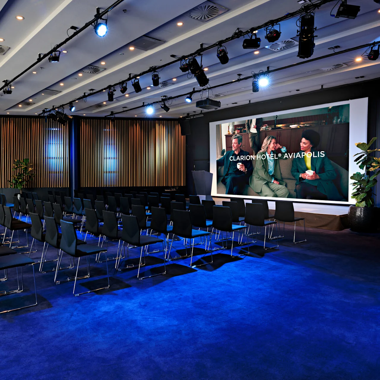 Rows of black chairs face a large screen displaying "Clarion Hotel® Aviapolis" with three people sitting and talking, in a conference room with blue carpet and wooden paneling.