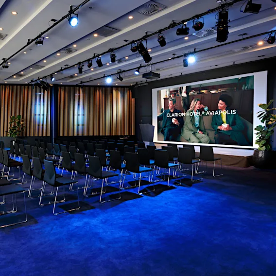 Rivit mustia tuoleja ovat suunnattu kohti suurta näyttöä, jossa lukee "Clarion Hotel® Aviapolis" ja kolme ihmistä istuu keskustelemassa, konferenssihuoneessa jossa on sininen matto ja puupanelointi.