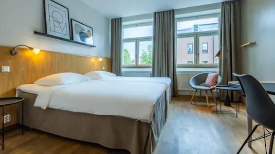 A neatly made bed in a modern hotel room with art on the wall, a sitting area, and a view of a city through the window.