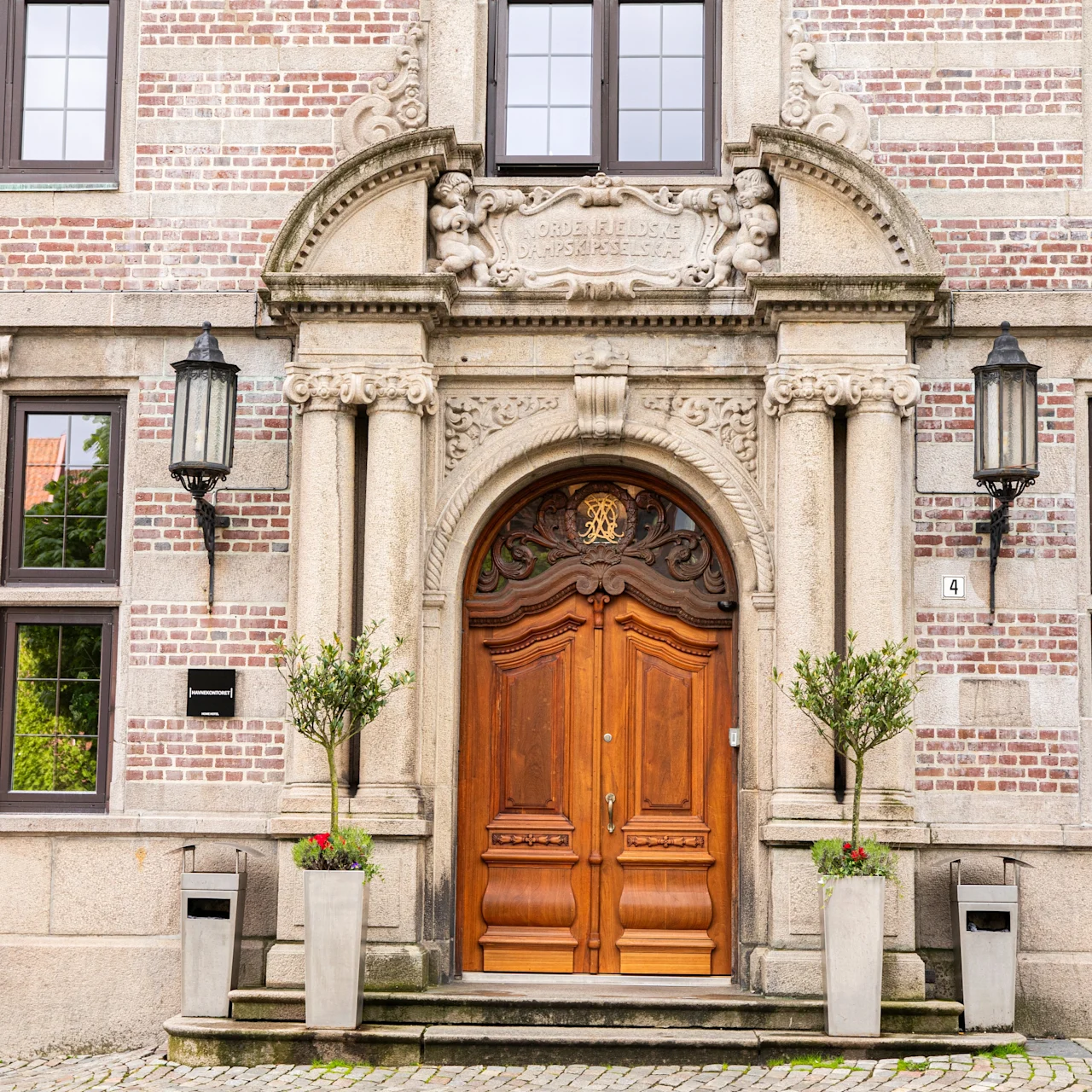 An ornate wooden door stands closed, surrounded by intricate stonework and two tall lanterns, flanked by potted plants and framed by multiple windows on a brick building. 